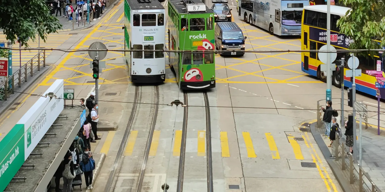 Two double-decker trams passing on Des Voeux Road Central in Hong Kong, with pedestrians waiting at the tram stop