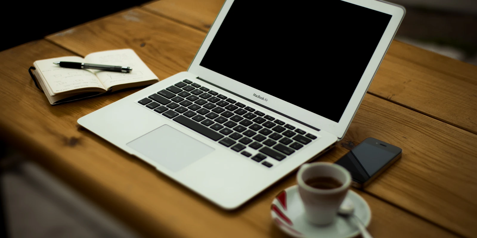 A laptop, smartphone, open notebook and coffee cup on a wooden table.